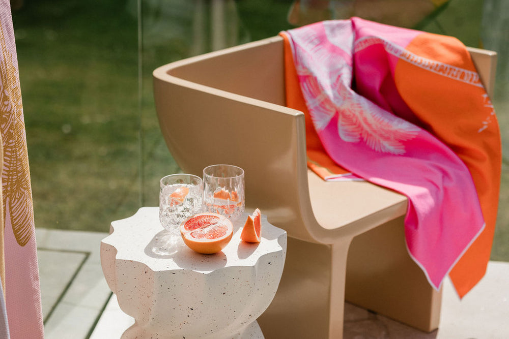 Bright orange and pink sand free towel hanging over the arm of a chair next to a large pool. Sand free towel has a hidden zipper corner pocket and a refreshing drink sitting on a table next to the pool.