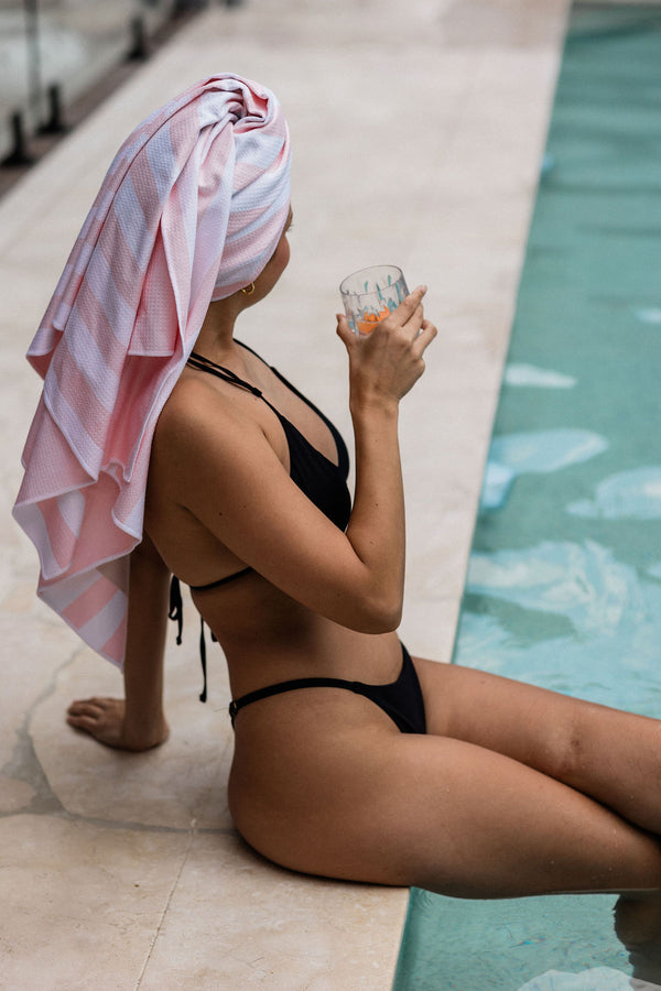 Woman, sitting with her feet in the pool, wearing a black string bikini. Her legs are folded, and she is drinking a refreshing orange drink. She has a luxurious pink and white striped towel wrapped around her hair.
